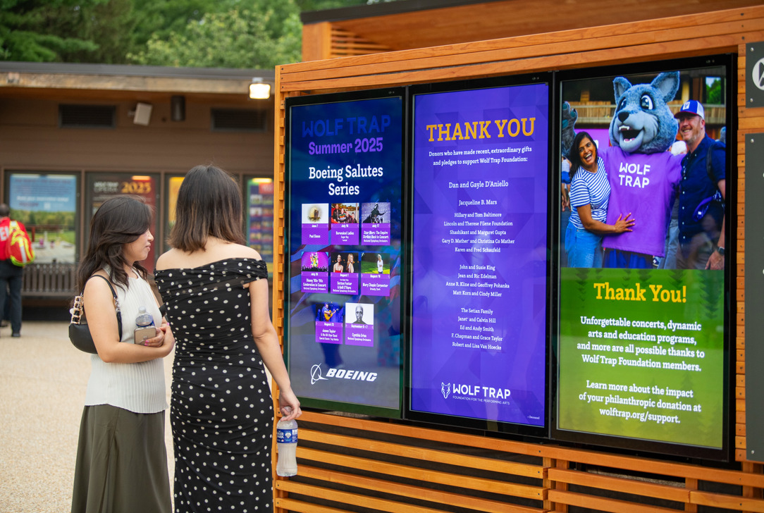 Two patrons on the Filene Center Plaza look at a Boeing performance series poster displayed on a screen.