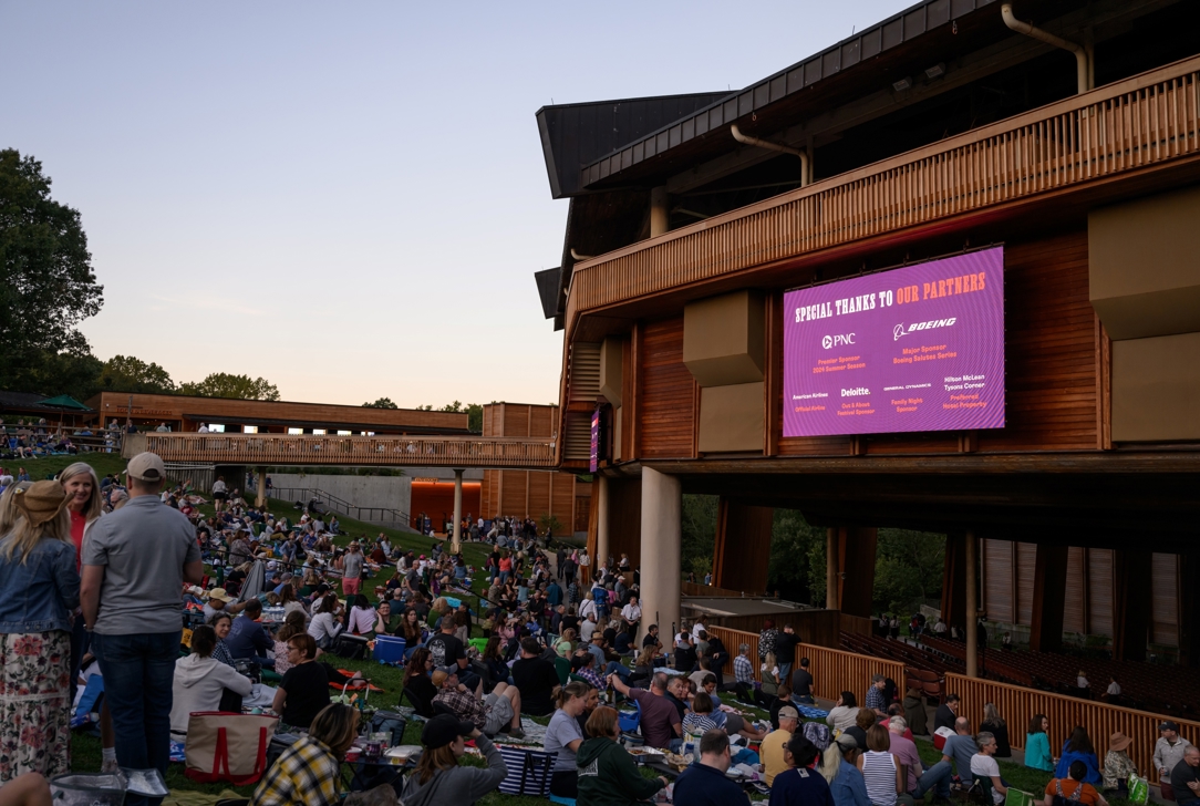 A list of corporate partners displayed on a screen above the lawn at Wolf Trap, with patrons picnicking below the screen.
